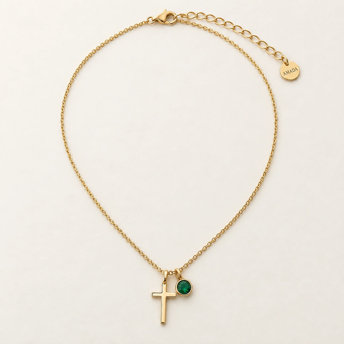 Amada Birthstone Cross Necklace in gold, shown with a green birthstone, on an ivory background.